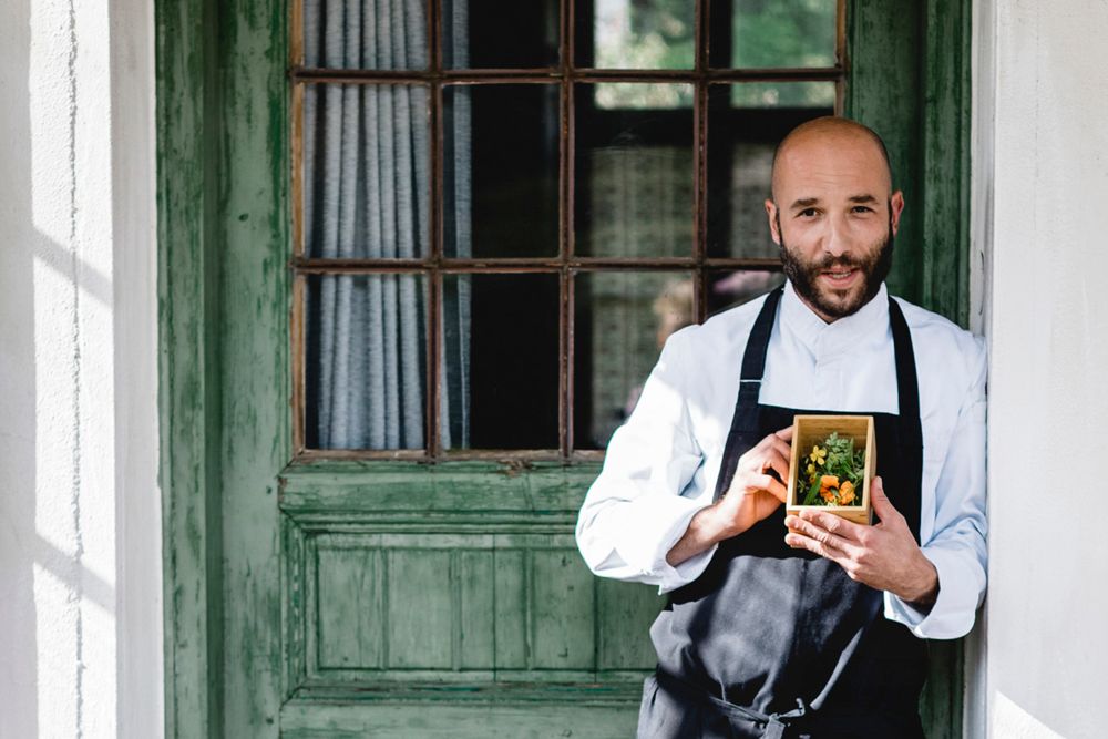 Ein Koch in weißer Uniform hält eine kleine Holzkiste mit Kräutern vor einer grünen Tür.