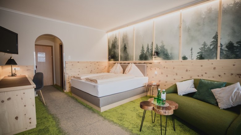 Ein gem&uuml;tliches Hotelzimmer mit Zirbenholzm&ouml;beln, einem Doppelbett, gr&uuml;nem Teppich und einem Sofa. An der Wand ein Bild von nebligem Wald.
