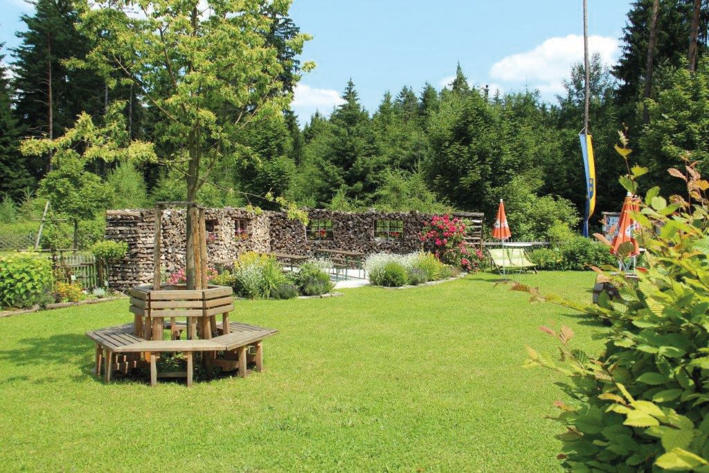 Ein grüner Garten mit Holzbänken um einen Baum, umgeben von Wald und Blumen.