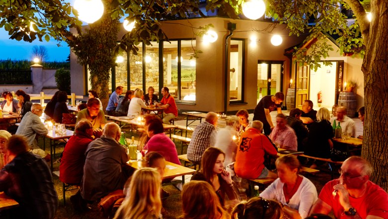 Menschen sitzen abends an Tischen im Freien, umgeben von B&auml;umen und Lichtern.