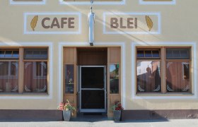 Eingang eines Cafés mit der Aufschrift 'Cafe Blei' und Blumendekoration.