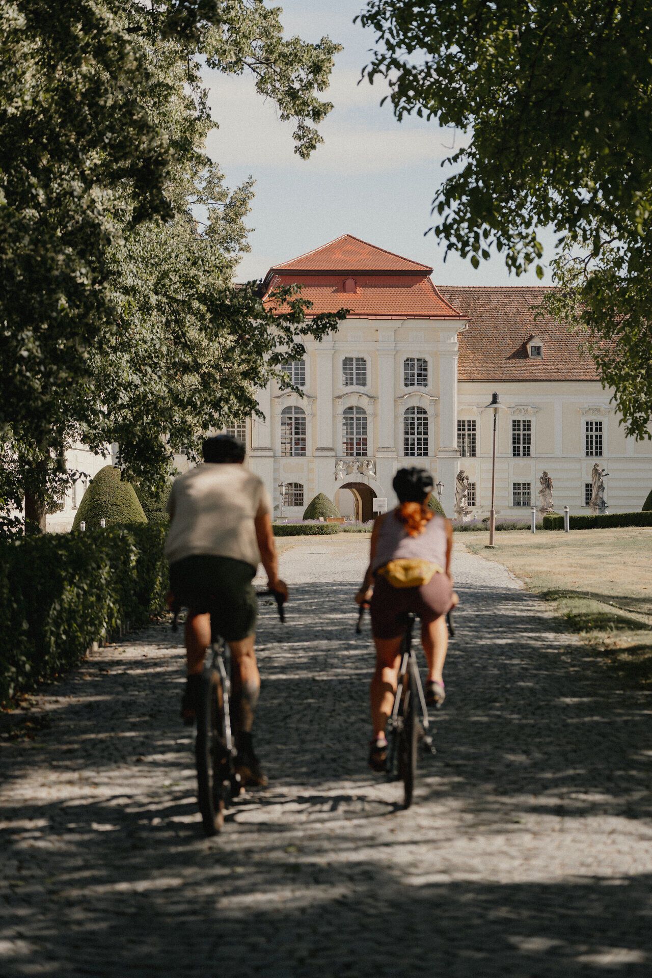 Zwei Radfahrer fahren auf einem gepflasterten Weg auf ein historisches Gebäude zu, umgeben von Bäumen.