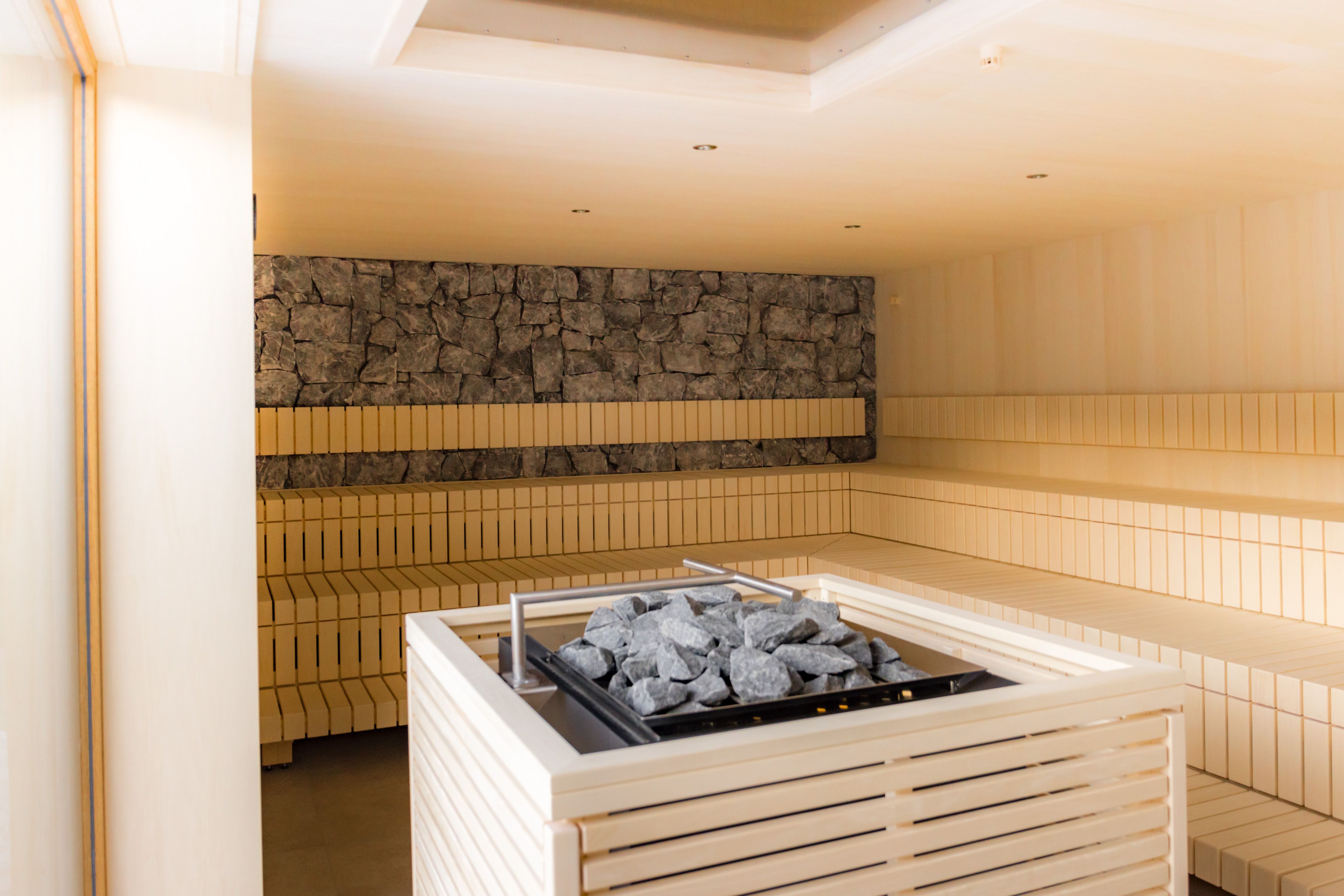 Innenansicht einer modernen Sauna mit Holzbänken und einem Ofen mit Steinen in der Mitte.