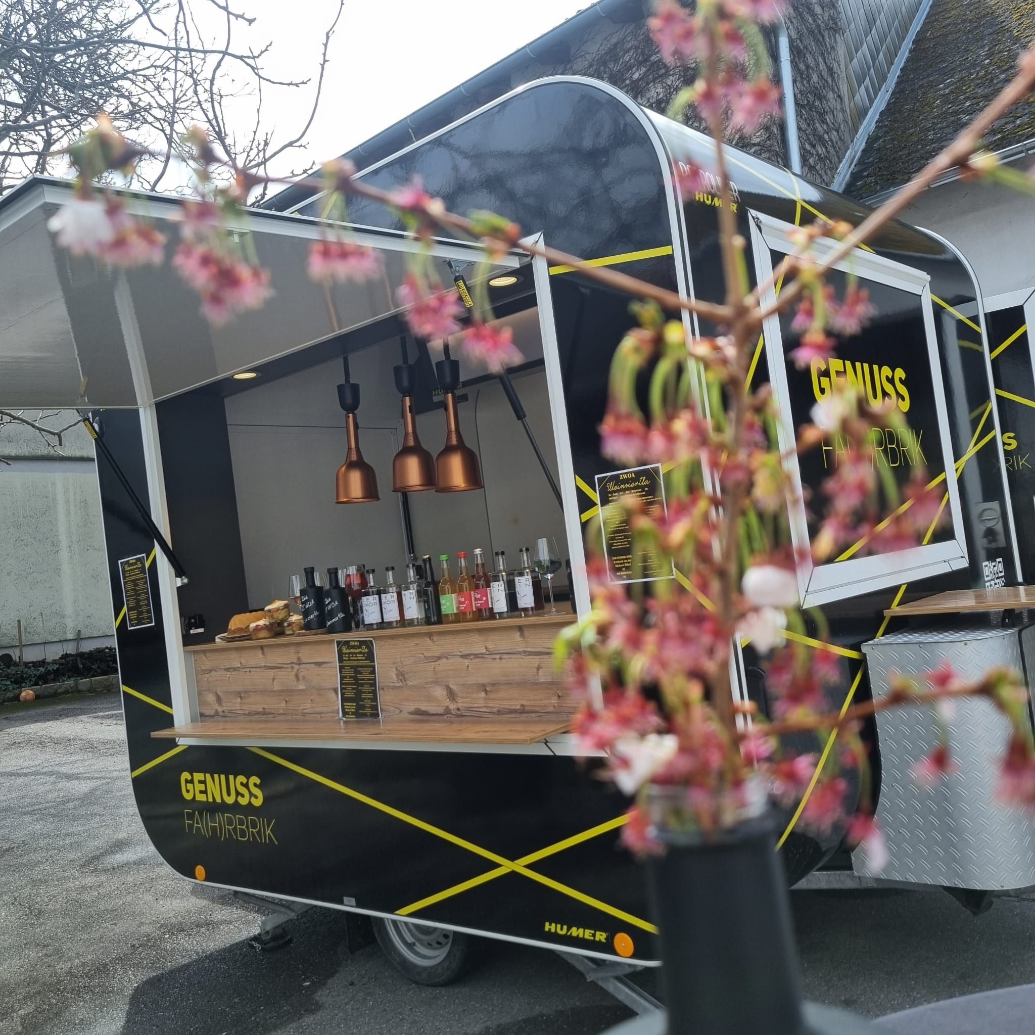 Ein schwarzer Foodtruck mit der Aufschrift 'Genuss Fahrbrik' steht im Freien. Im Vordergrund sind unscharfe Blüten zu sehen.
