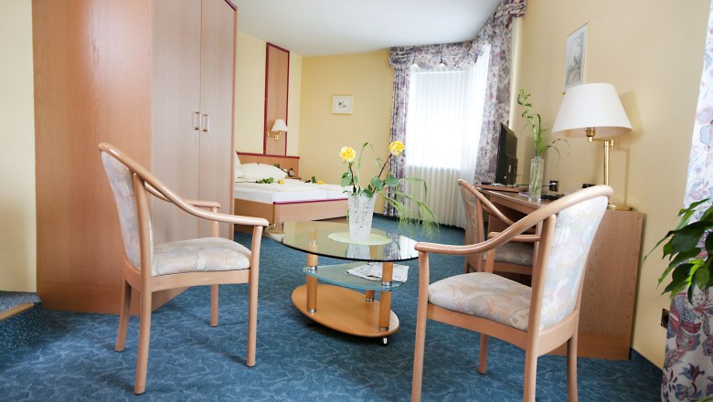 Hotelzimmer mit Doppelbett, zwei Stühlen, Glastisch und TV.