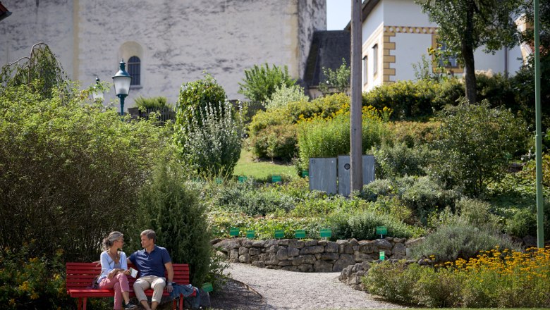 Ein Paar sitzt auf einer roten Bank in einem Garten vor der Wehrkirche Bad Schönau.