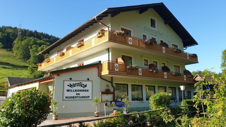 Ein traditionelles Gasthaus namens Hubertushof mit Blumen auf den Balkonen, umgeben von gr&uuml;ner Landschaft und blauem Himmel.