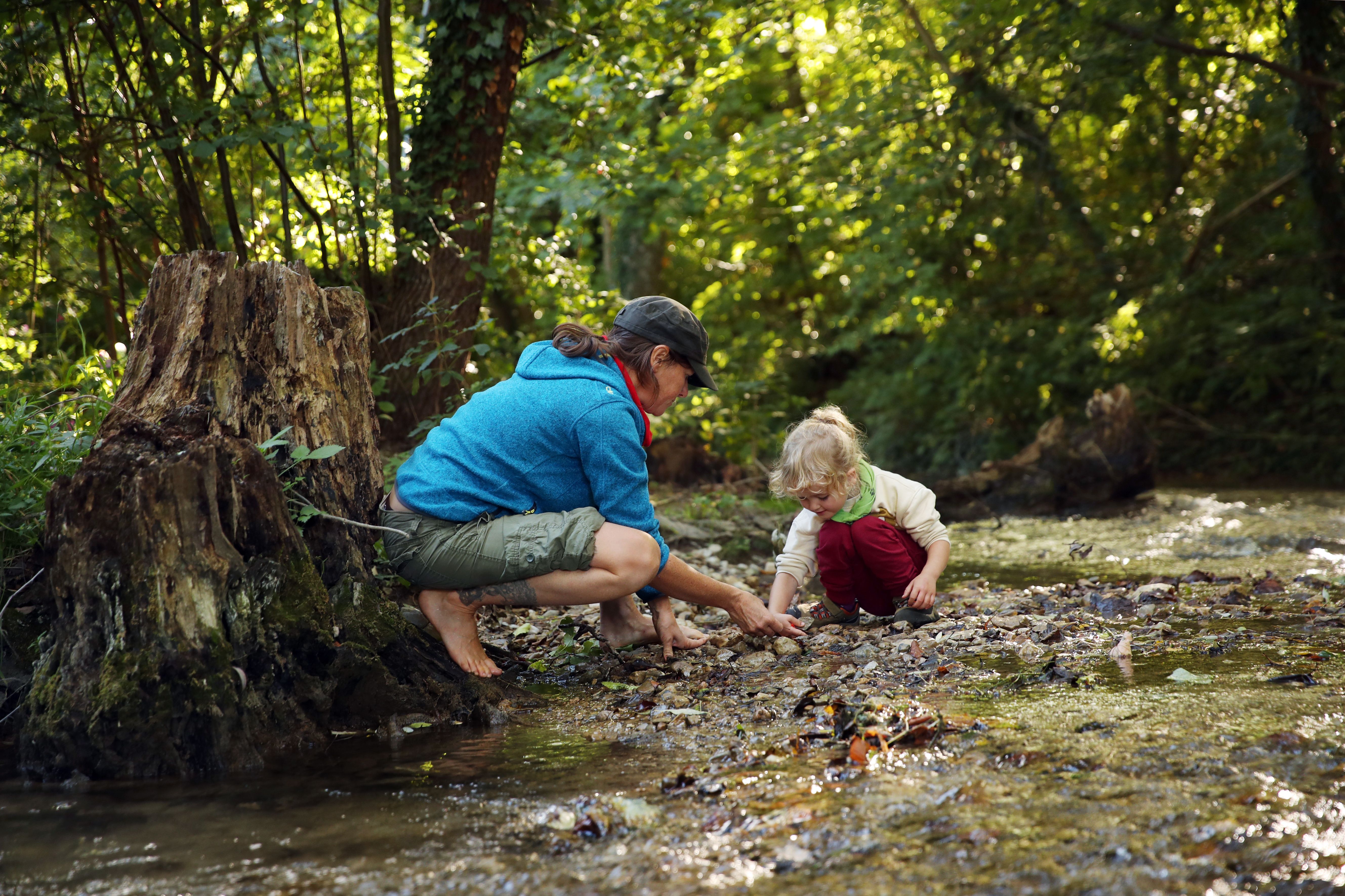 Eine erwachsene Person und ein Kind spielen an einem Bach in einem Wald.