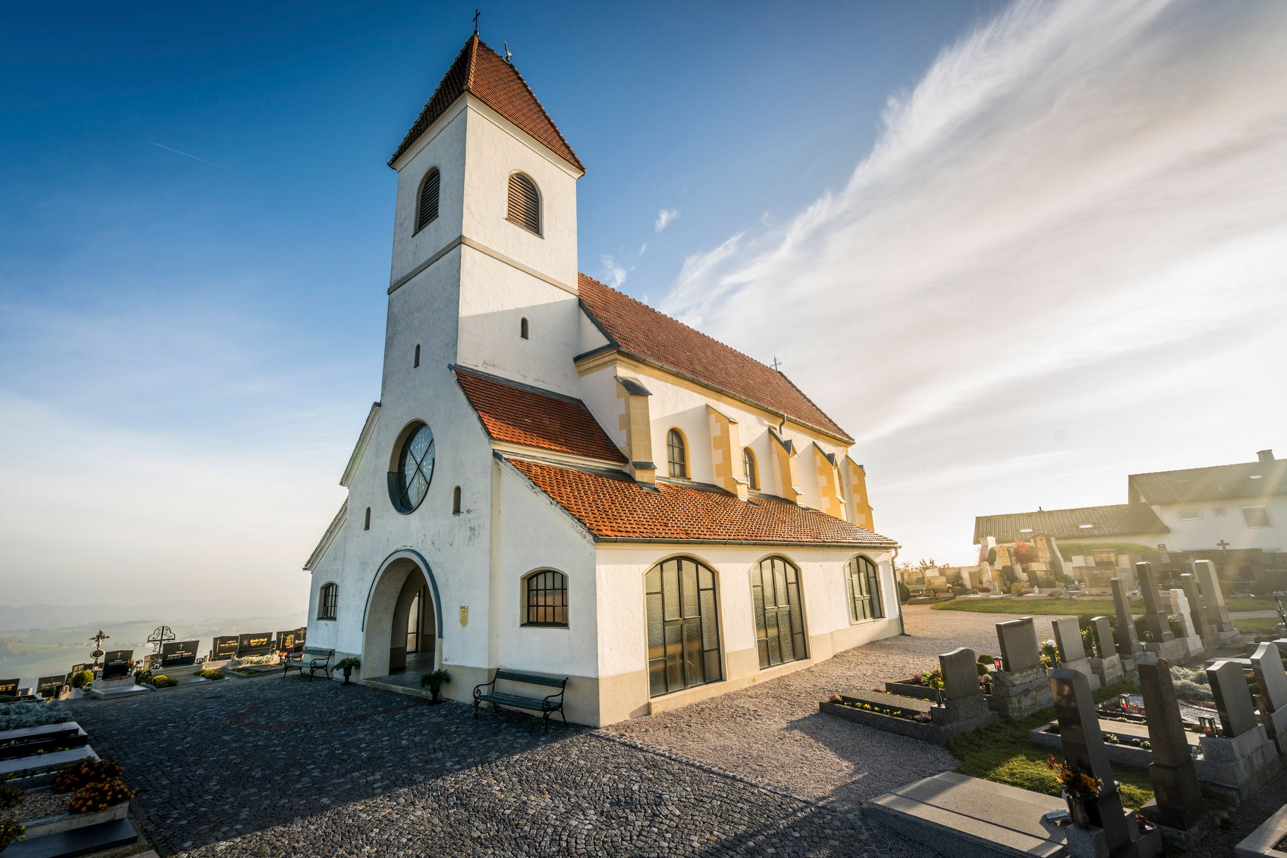 Eine weiße Kirche mit rotem Ziegeldach und einem Friedhof im Vordergrund bei sonnigem Wetter.
