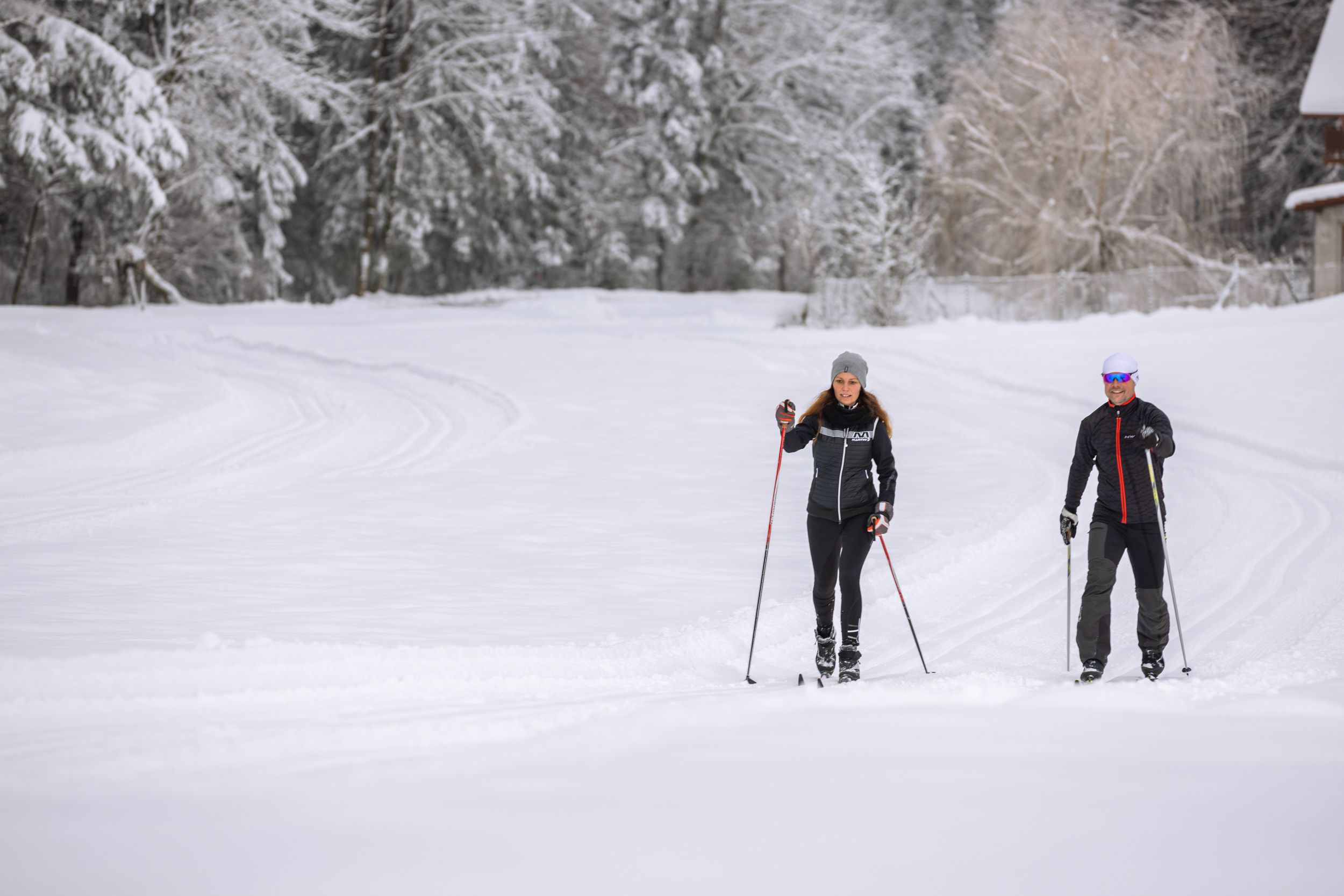 Zwei Personen beim Langlaufen auf einer verschneiten Loipe, umgeben von schneebedeckten Bäumen.