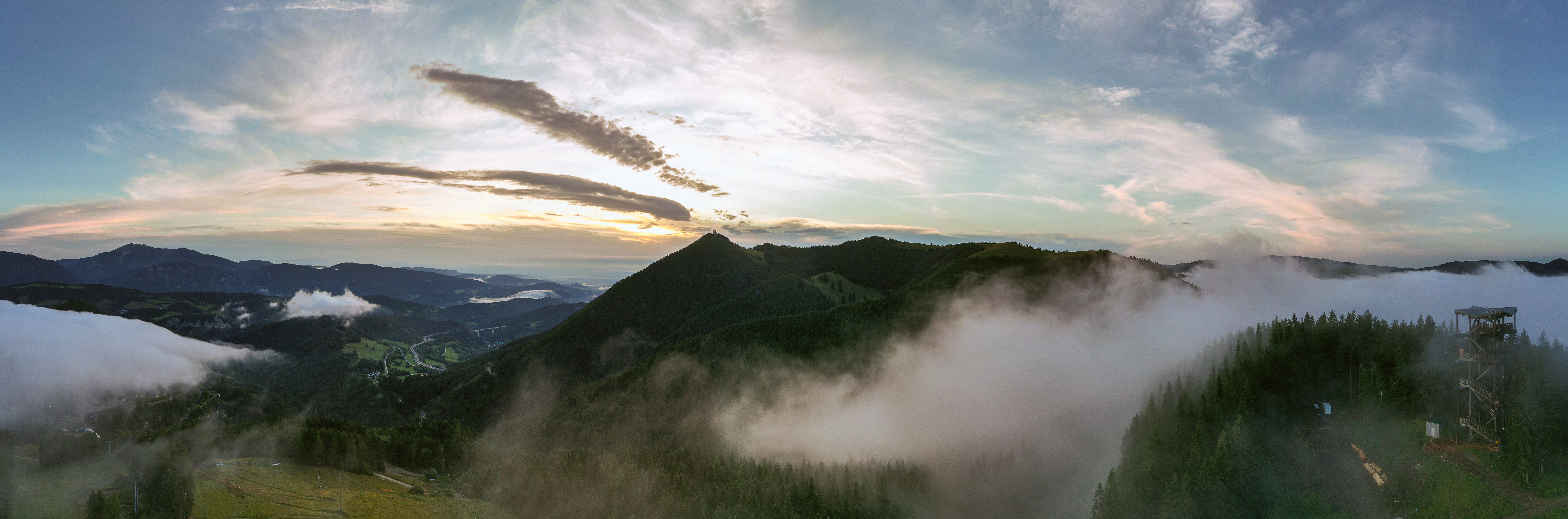 Panoramablick auf den Sonnwendstein bei Sonnenaufgang mit Nebel und einem Aussichtsturm im Vordergrund.