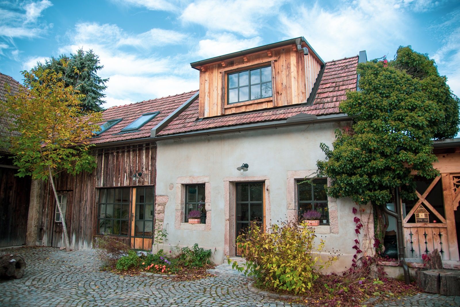 Rustikales Haus mit Ziegeldach und Holzverkleidung, umgeben von Pflanzen und Kopfsteinpflaster.