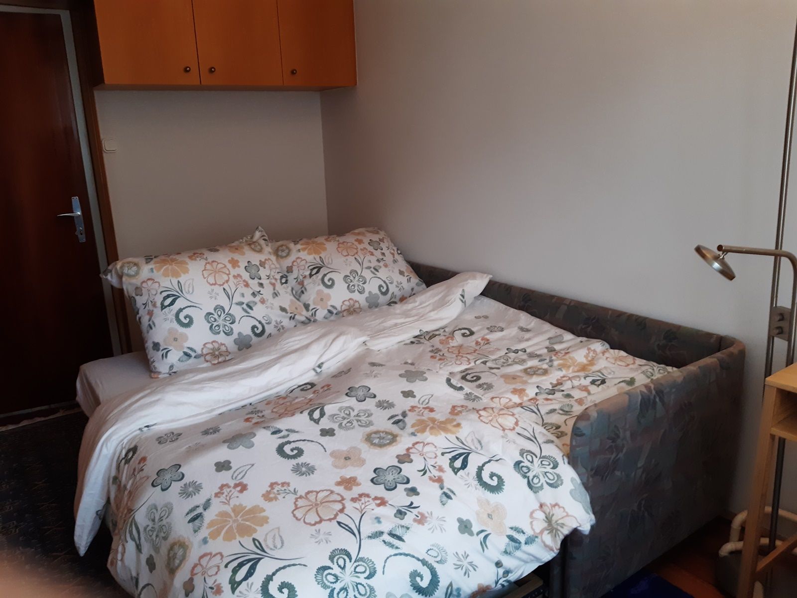 Ein kleines Zimmer mit einem ausziehbaren Sofa, das als Bett für zwei Personen dient, mit floraler Bettwäsche und einem Leselicht daneben.