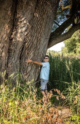 Umgeben von üppigem Grün und der majestätischen Präsenz eines alten Baumes, strahlt die Szenerie eine friedliche Sommeratmosphäre aus. Ein Abenteurer umarmt den Stamm, während die sanften Wellen des Wassers in der Nähe leise plätschern. Diese idyllische Umgebung lädt dazu ein, die Natur in vollen Zügen zu genießen.