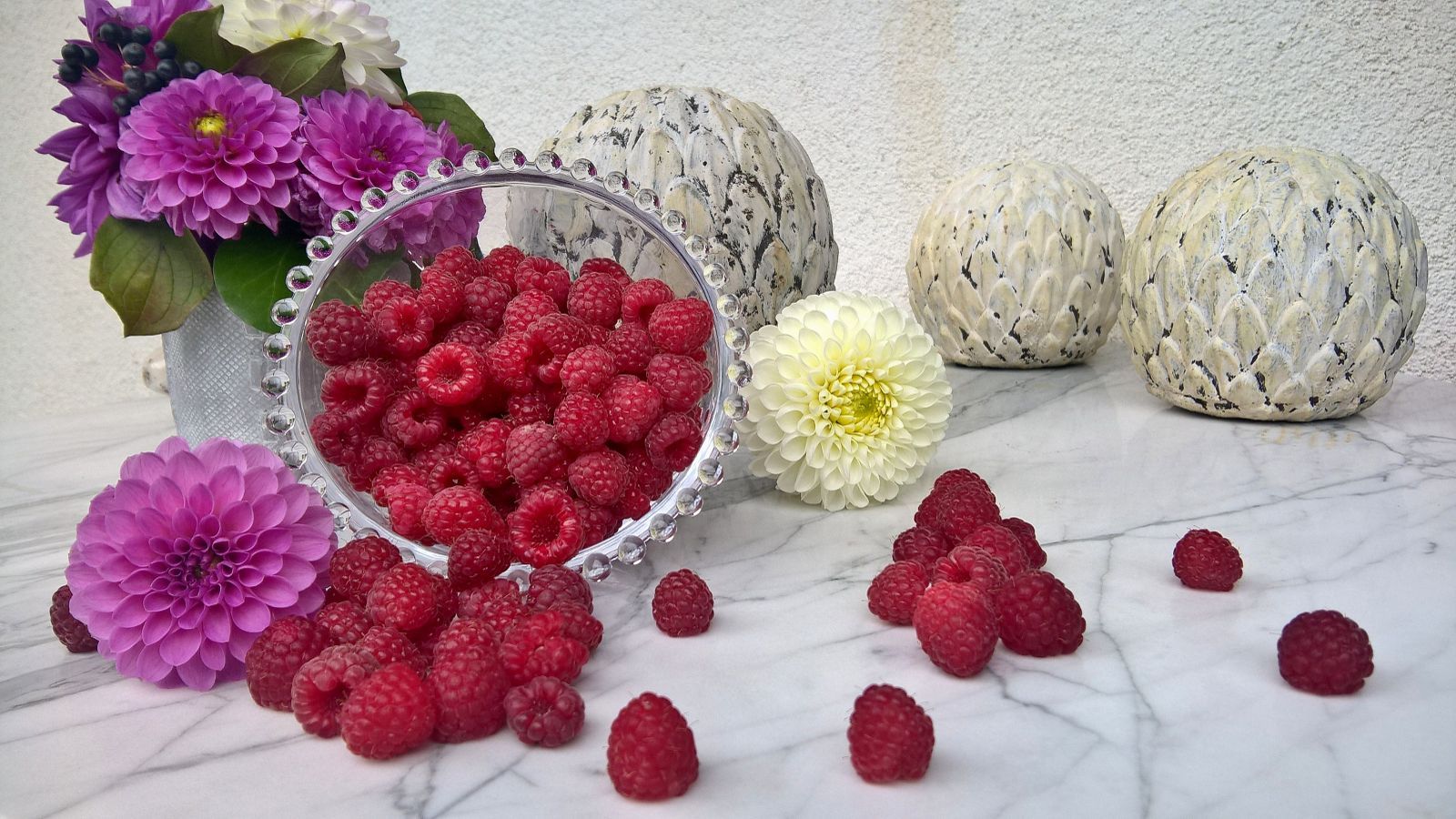 Himbeeren in einer Schale mit Blumen und Dekokugeln auf einem Marmortisch.
