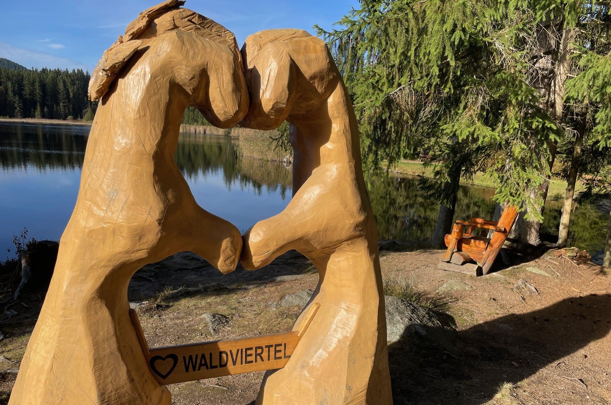 Holzskulptur von zwei Händen, die ein Herz formen, mit der Aufschrift 'Waldviertel' vor einem See und Wald.