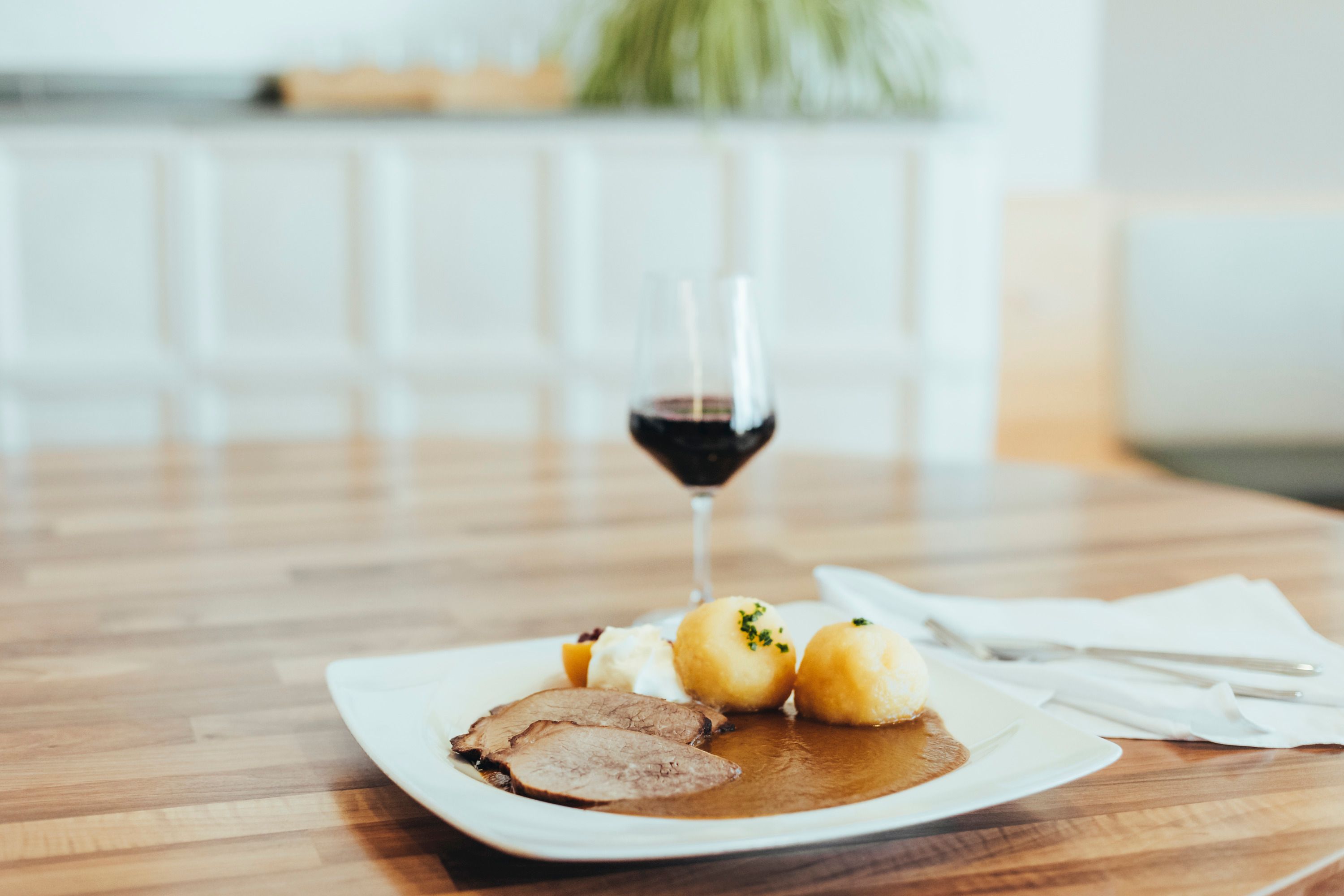 Ein Teller mit Rindsbraten, Knödeln und Soße auf einem Holztisch, daneben ein Glas Rotwein.