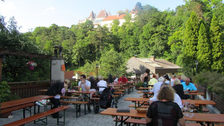 Menschen sitzen an Holztischen im Freien mit Blick auf ein Schloss im Hintergrund.