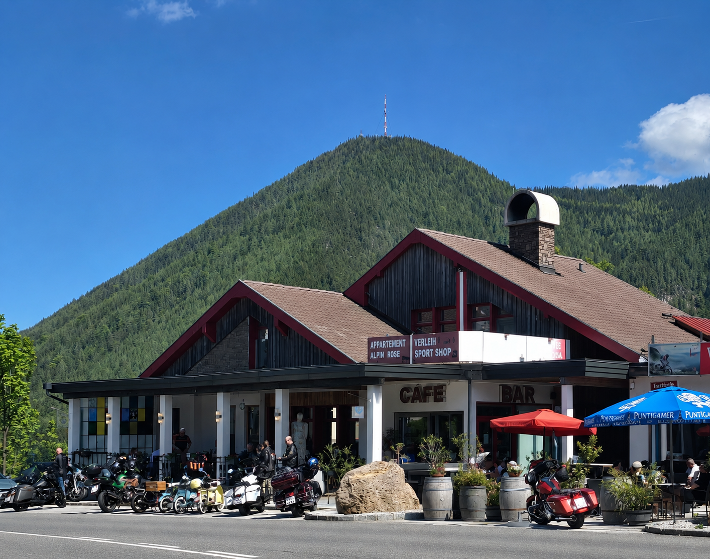Ein Café und Bar mit Motorrädern davor, umgeben von Bergen und Bäumen.