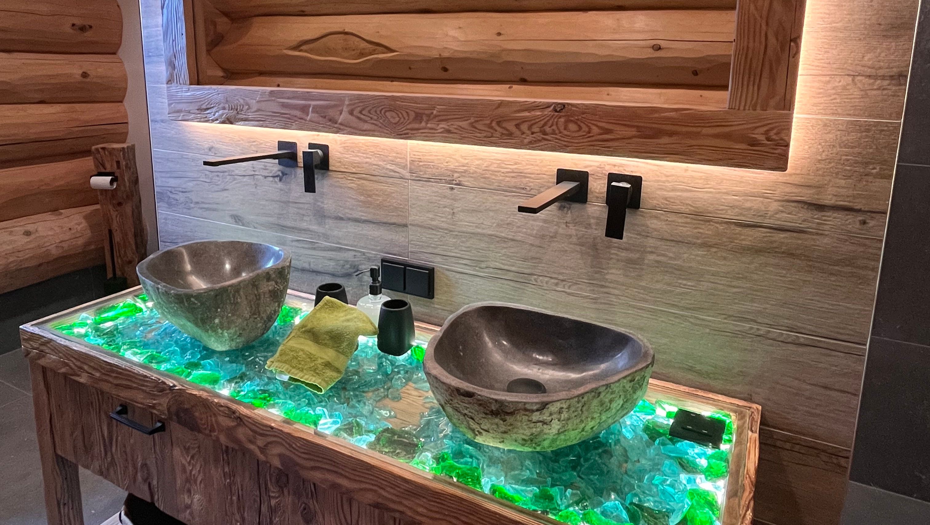 Modernes Badezimmer mit zwei steinernen Waschbecken auf einem beleuchteten Glastisch.