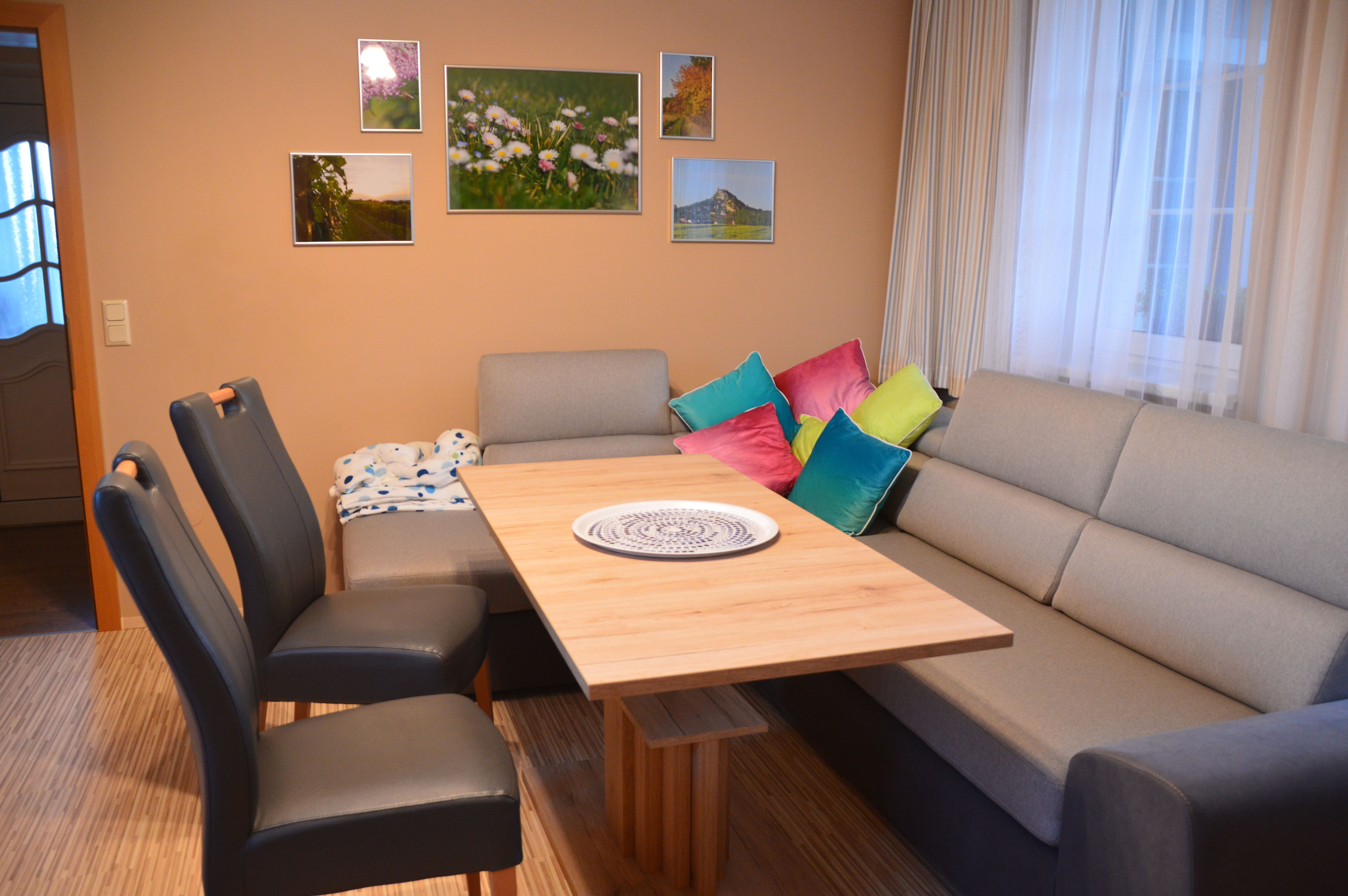 Wohn- und Essbereich mit grauem Sofa, Holztisch, bunten Kissen und Wandbildern.
