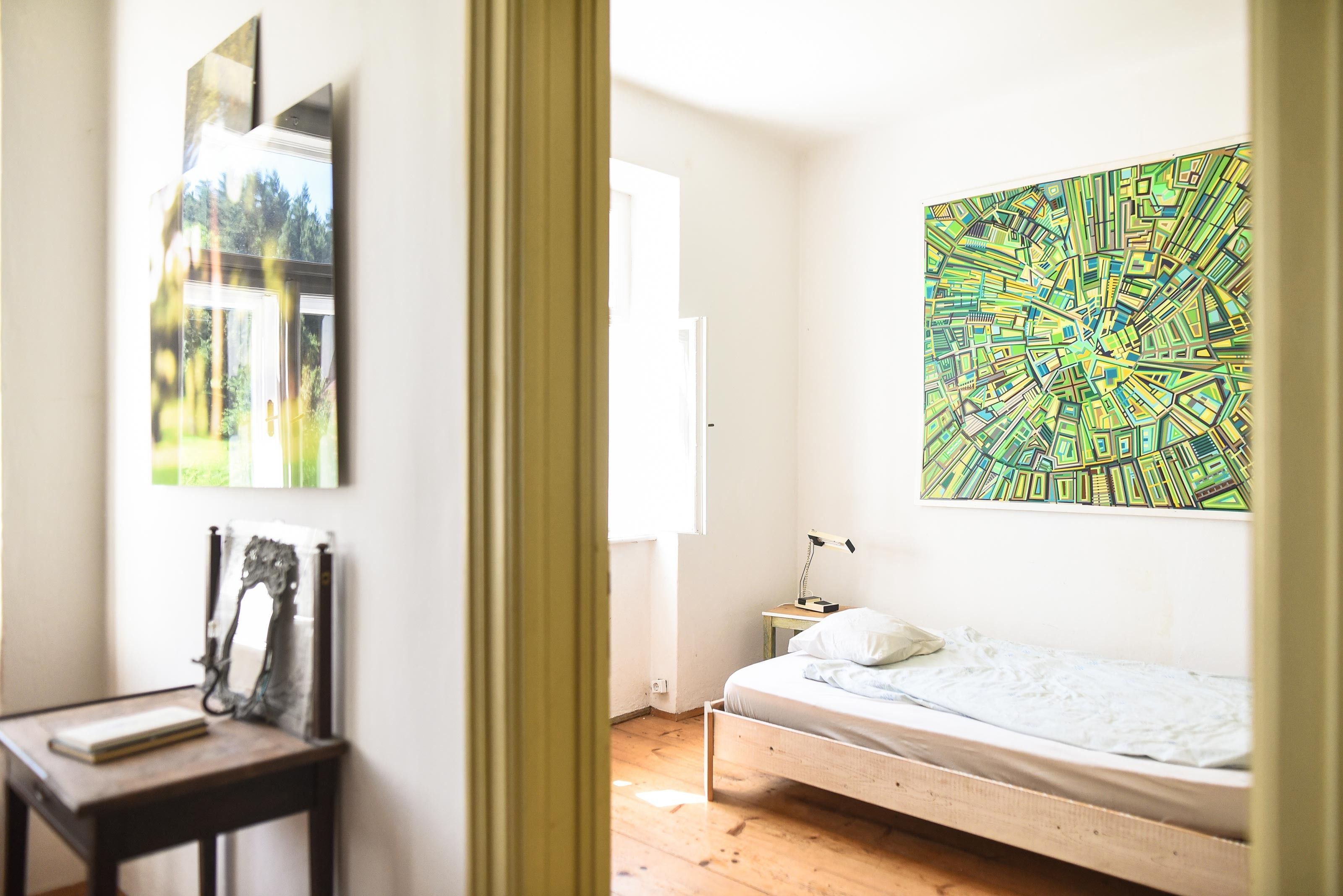 Ein einfach eingerichtetes Schlafzimmer mit einem Einzelbett, einem kleinen Tisch und einem großen, bunten Gemälde an der Wand.