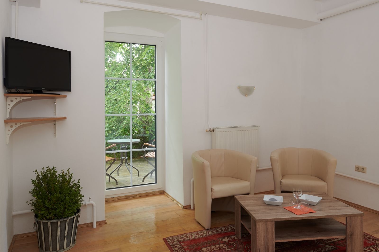 Wohnzimmer mit zwei beigen Sesseln, Holztisch, Fernseher und Balkon mit Tisch und Stühlen.