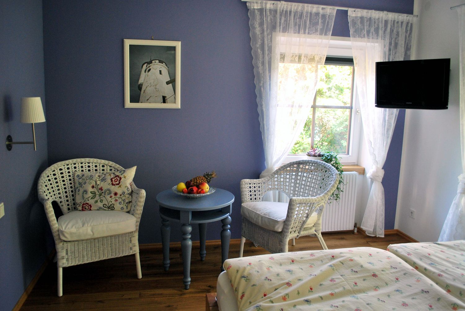 Gemütliches Zimmer mit blauen Wänden, zwei Korbsesseln, einem runden Tisch mit Obstschale und einem Fenster mit weißen Vorhängen.