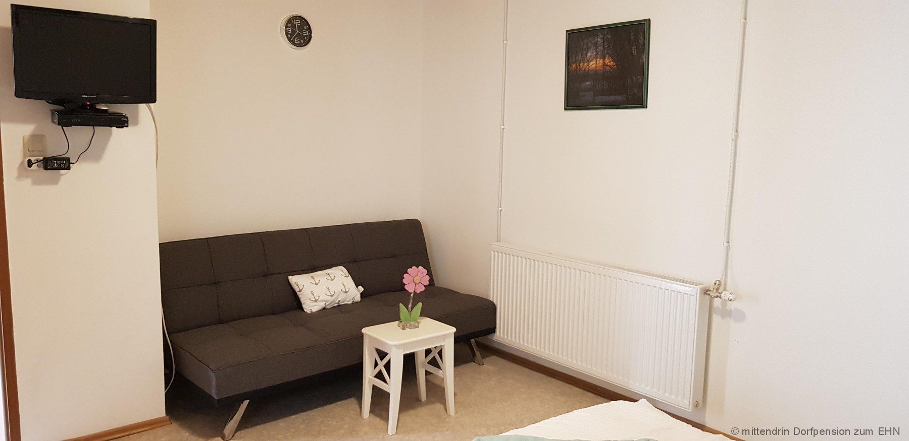 Fernseher an der Wand, daneben ein graues Sitzsofa und einem kleinem Tisch.