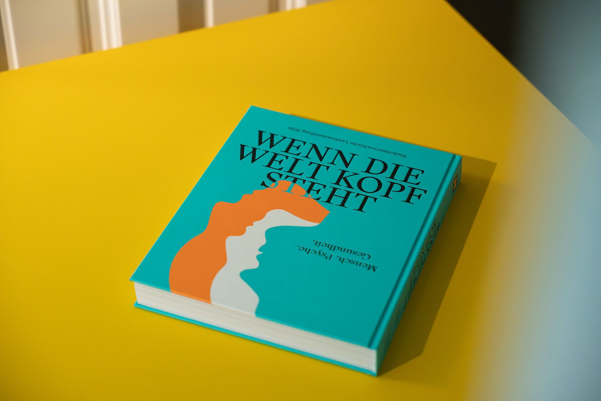 Buch „Wenn die Welt Kopf steht – Mensch. Psyche. Gesundheit.“ der Niederösterreichischen Landesausstellung 2026 auf gelbem Tisch, mit grafischem Cover in Türkis und Orange.
