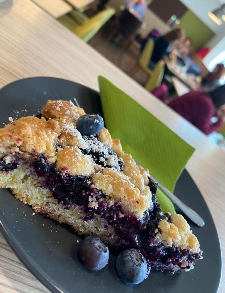 Ein Stück Heidelbeerkuchen auf einem schwarzen Teller mit frischen Heidelbeeren und Puderzucker, im Hintergrund unscharfe Café-Szene.