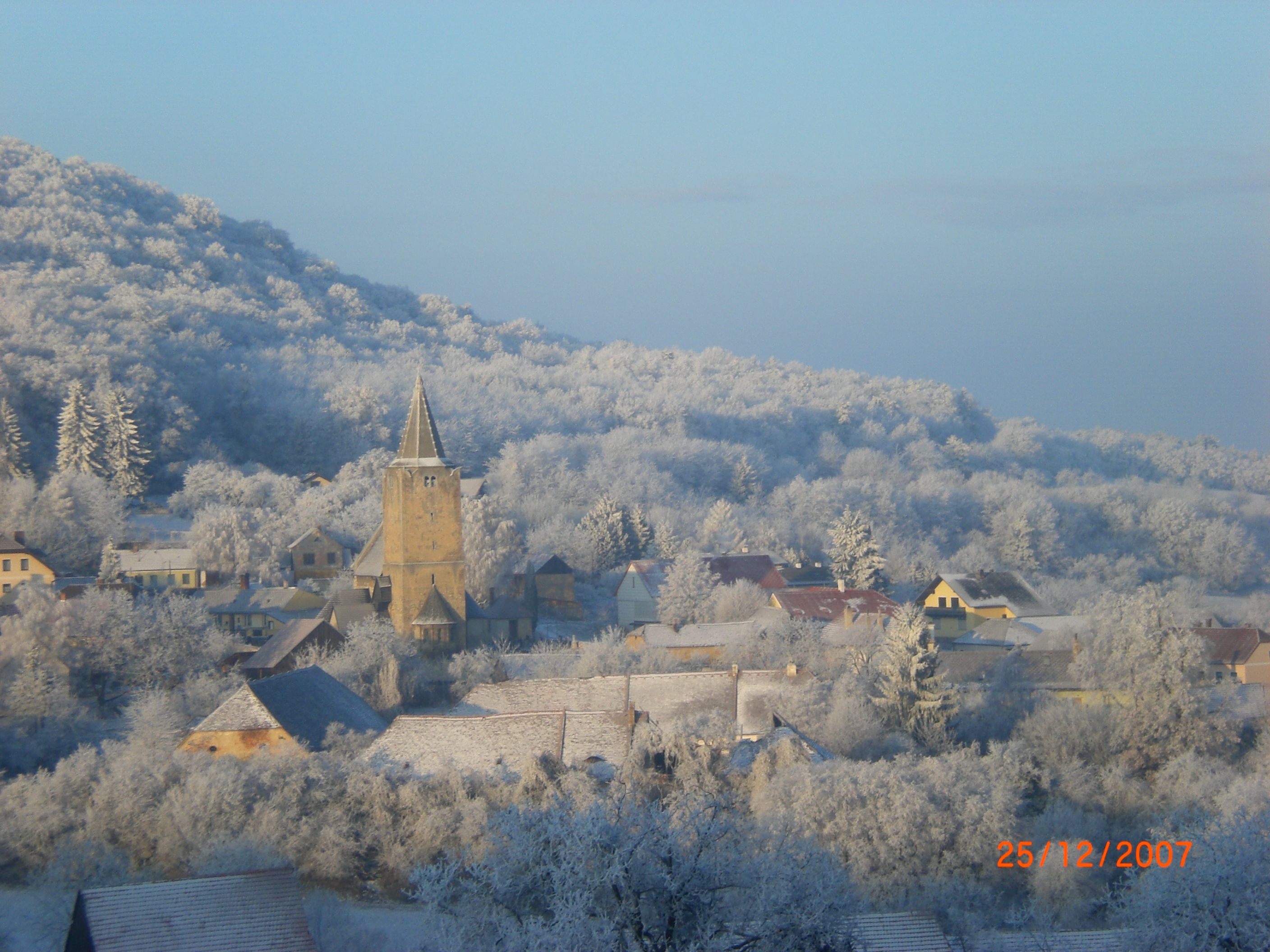Winterlandschaft mit Dorf und Kirche, umgeben von schneebedeckten Bäumen.