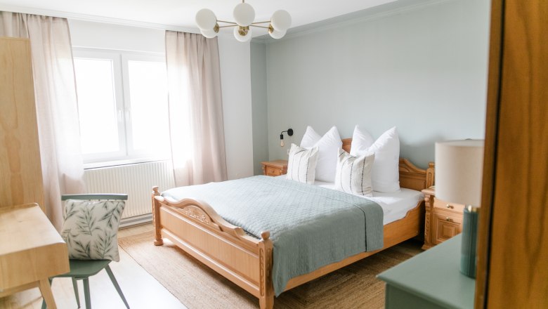 Ein gemütliches Schlafzimmer mit einem Holzbett, blauer Bettdecke, Nachttischen und einem Schreibtisch mit Stuhl. Helle Vorhänge und ein Fenster sind zu sehen.