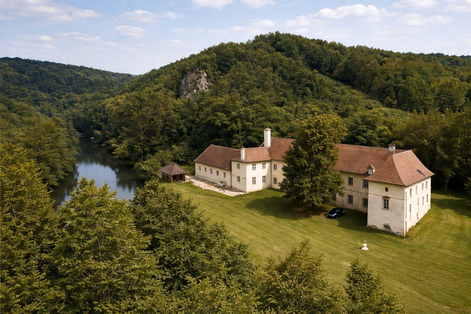 Luftaufnahme eines großen, historischen Gebäudes mit rotem Dach, umgeben von Wald und einem Fluss.