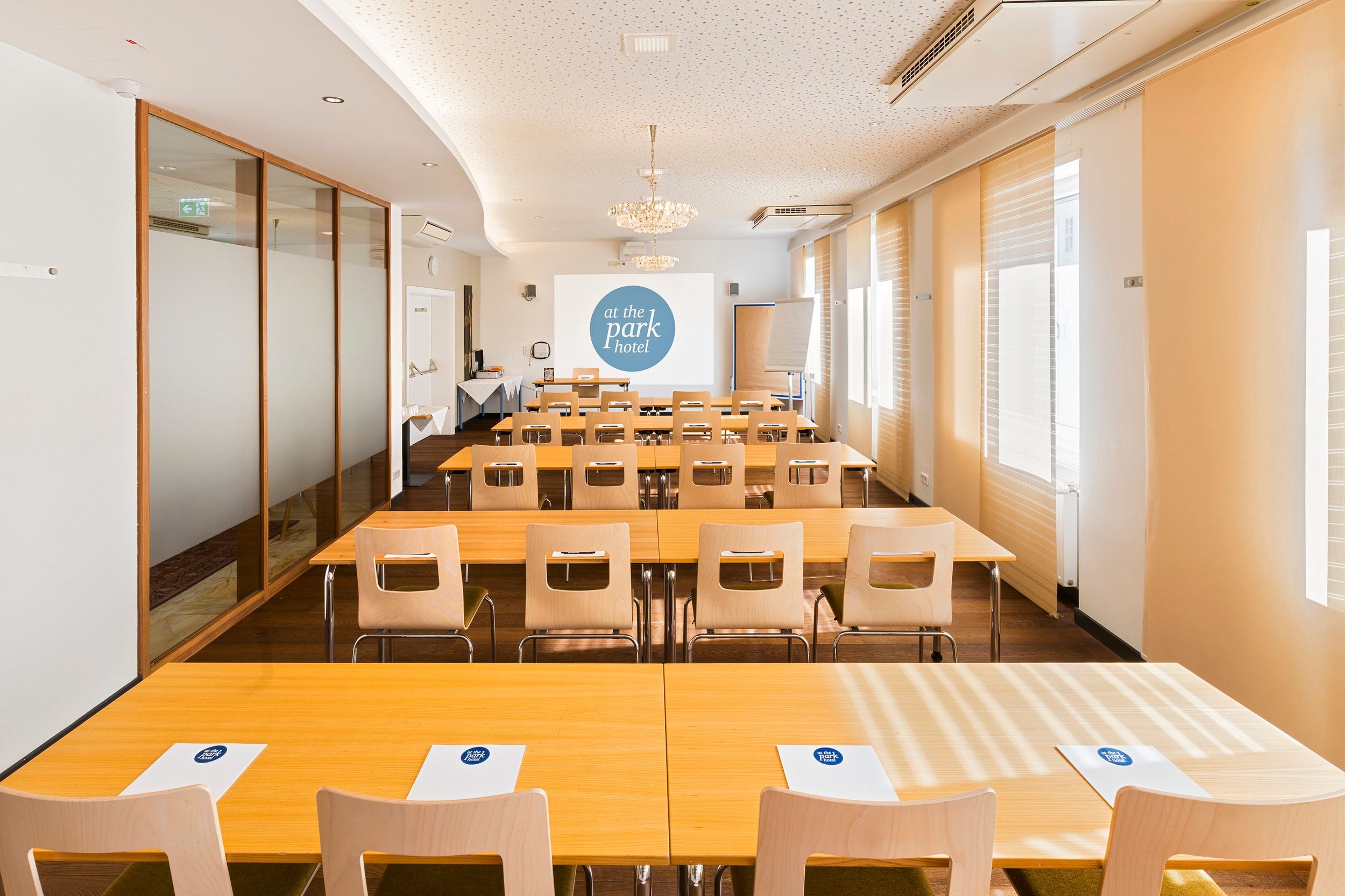 Ein Konferenzraum mit Tischen und Stühlen, einem Projektorbildschirm und Notizblöcken mit dem Logo "at the park hotel".