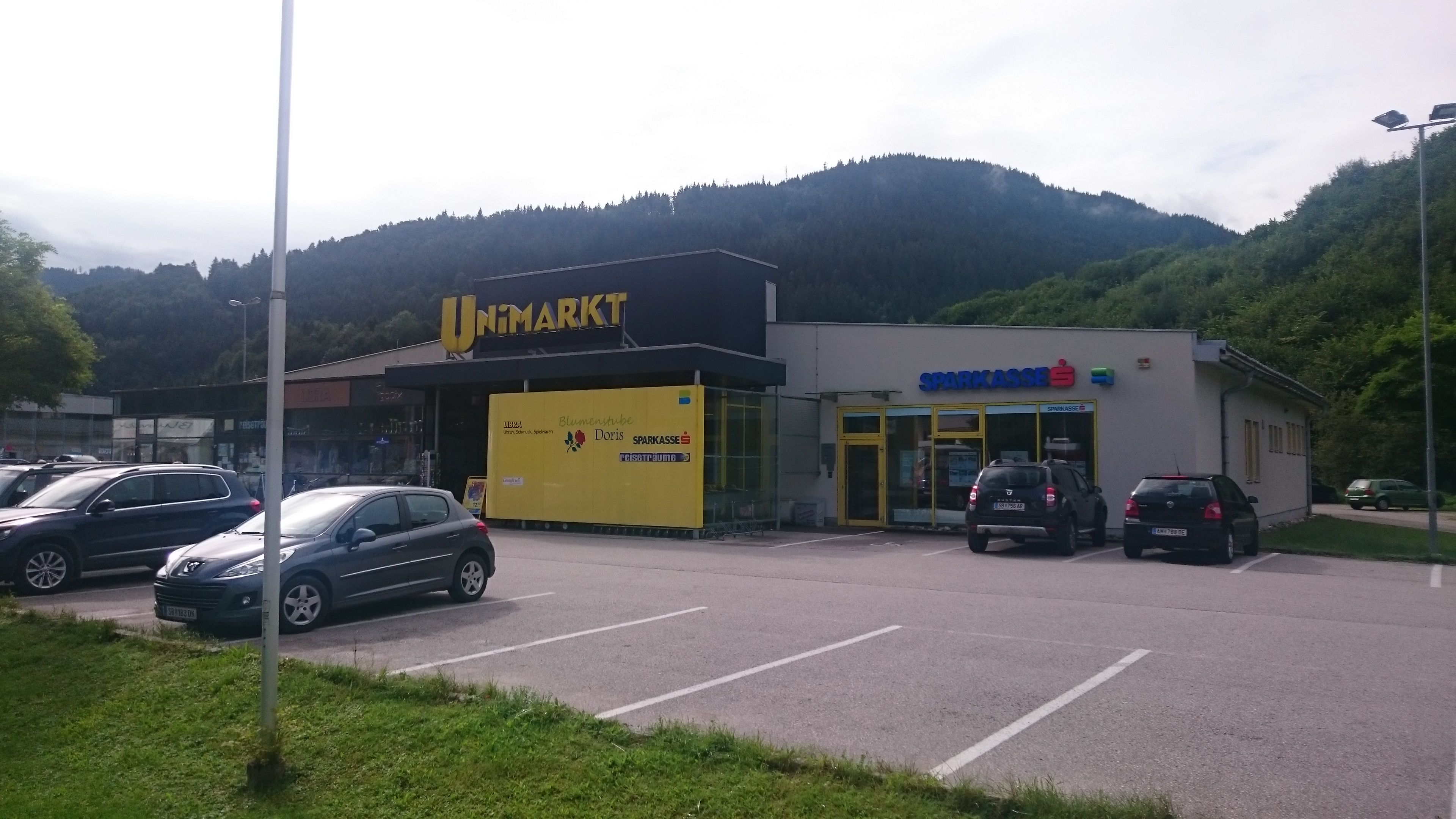 Einkaufszentrum mit Unimarkt und Sparkasse vor bewaldeten Hügeln.