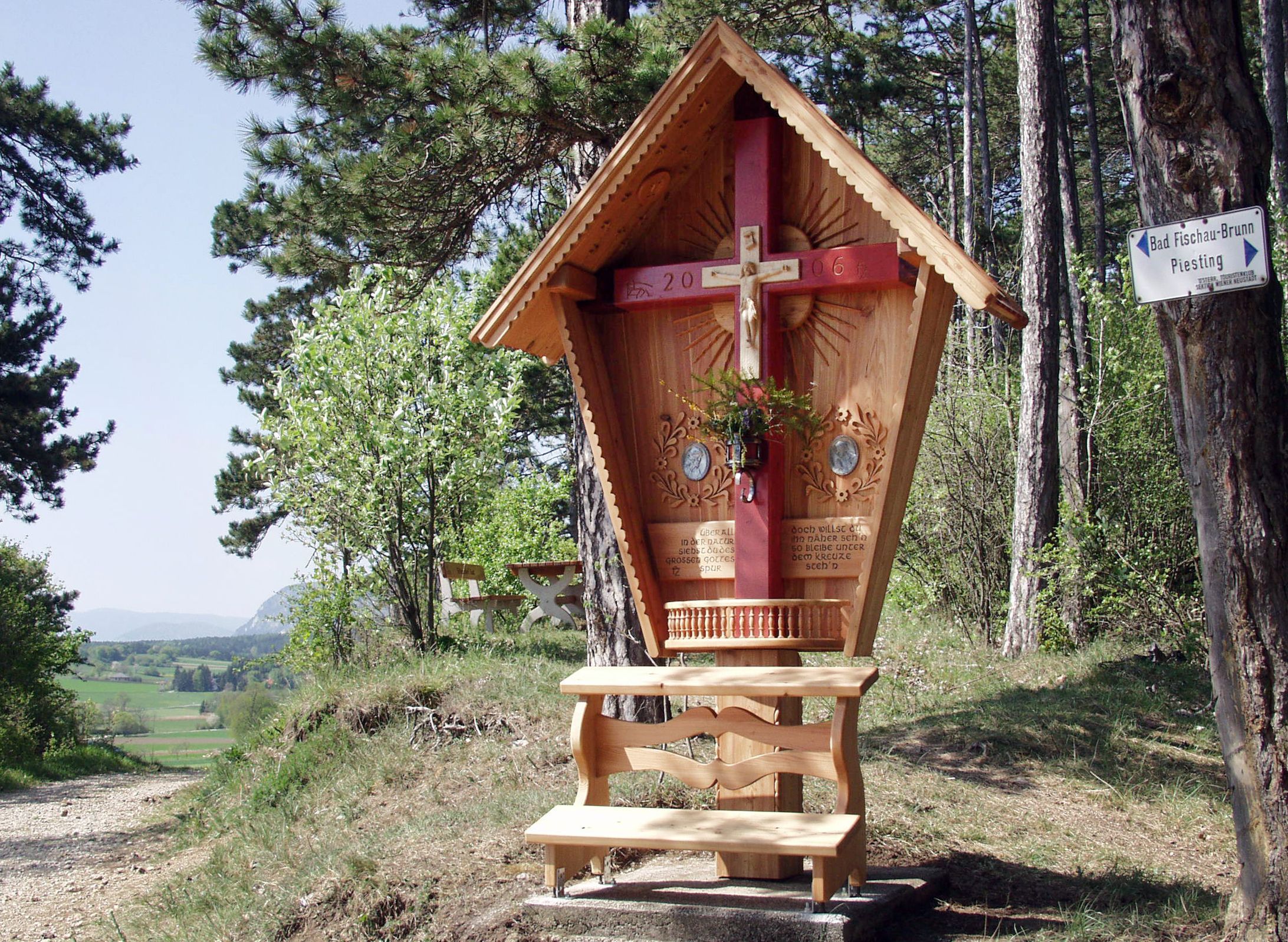 Holzkreuz mit rotem Balken und geschnitztem Dach in einer Waldlandschaft.