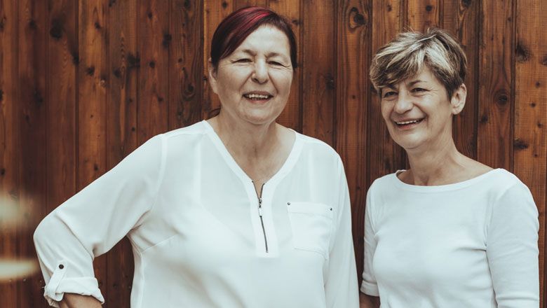 Zwei Frauen in weißen Blusen stehen vor einer Holzwand und lächeln.