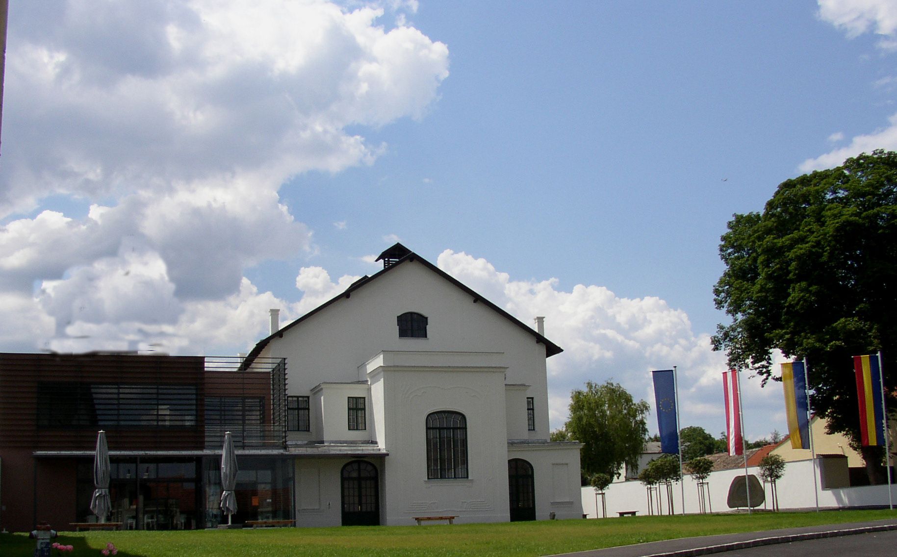 Ein modernes Konzerthaus mit weißen Wänden und großen Fenstern, umgeben von Flaggen und Bäumen, unter einem bewölkten Himmel.