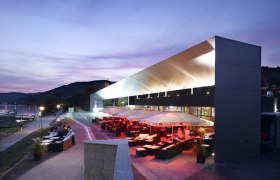 Modernes Gebäude am Donau-Ufer bei Dämmerung mit beleuchteter Terrasse und Sitzbereichen.