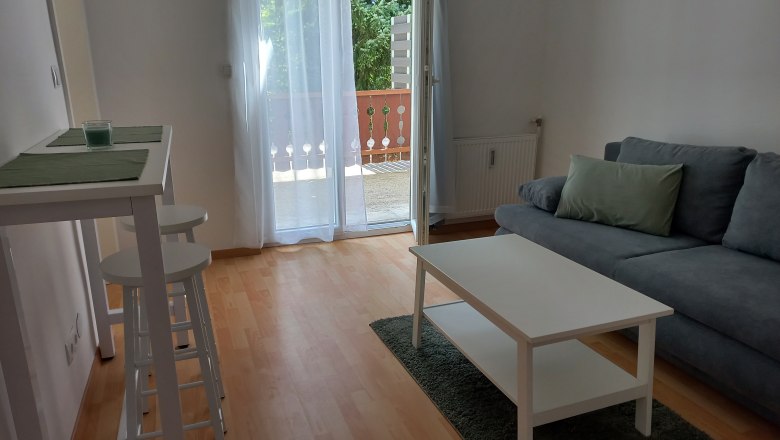 Modernes Wohnzimmer mit grauem Sofa, weißem Couchtisch und Barhockern. Balkon mit Glas-Schiebetür im Hintergrund.