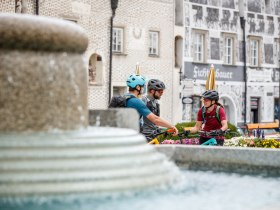 Drei Radfahrer genie&szlig;en eine entspannte Pause am Stadtplatz, umgeben von bl&uuml;henden Blumen und der charmanten Architektur. Die Atmosph&auml;re ist lebhaft und einladend, w&auml;hrend die frische Luft und die Natur zum Erkunden einladen.