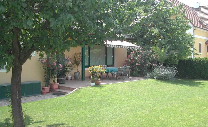 Ein gepflegter Garten mit Rasen, Bäumen und einer Terrasse vor einem gelben Haus.