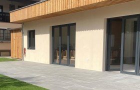 Moderne Gebäudeaußenansicht mit Holzverkleidung und großen Fenstern, umgeben von einer Terrasse und Rasenfläche.