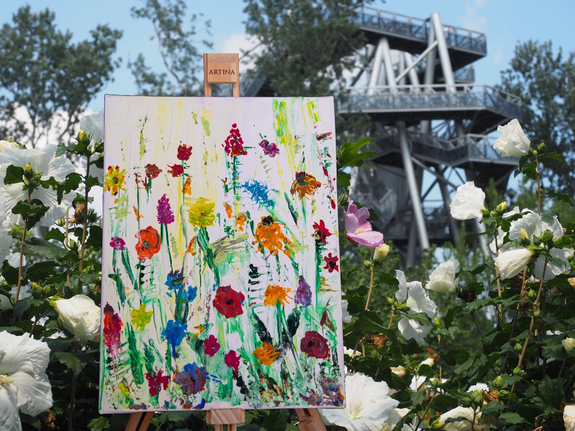 Ein Gemälde mit bunten Blumen steht auf einer Staffelei in einem Garten, umgeben von weißen Blüten. Im Hintergrund ist ein Aussichtsturm zu sehen.