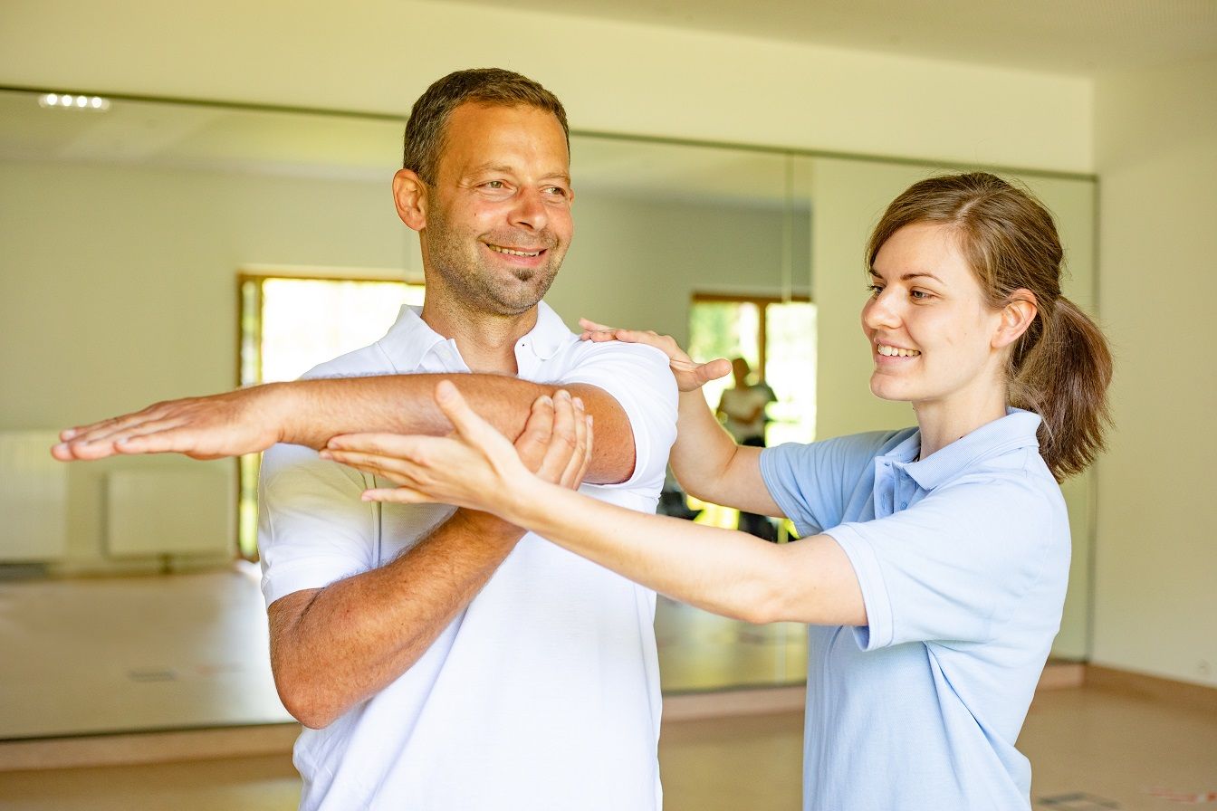 Ein Physiotherapeut hilft einem Mann bei einer Armübung in einem hellen Raum mit Spiegeln.