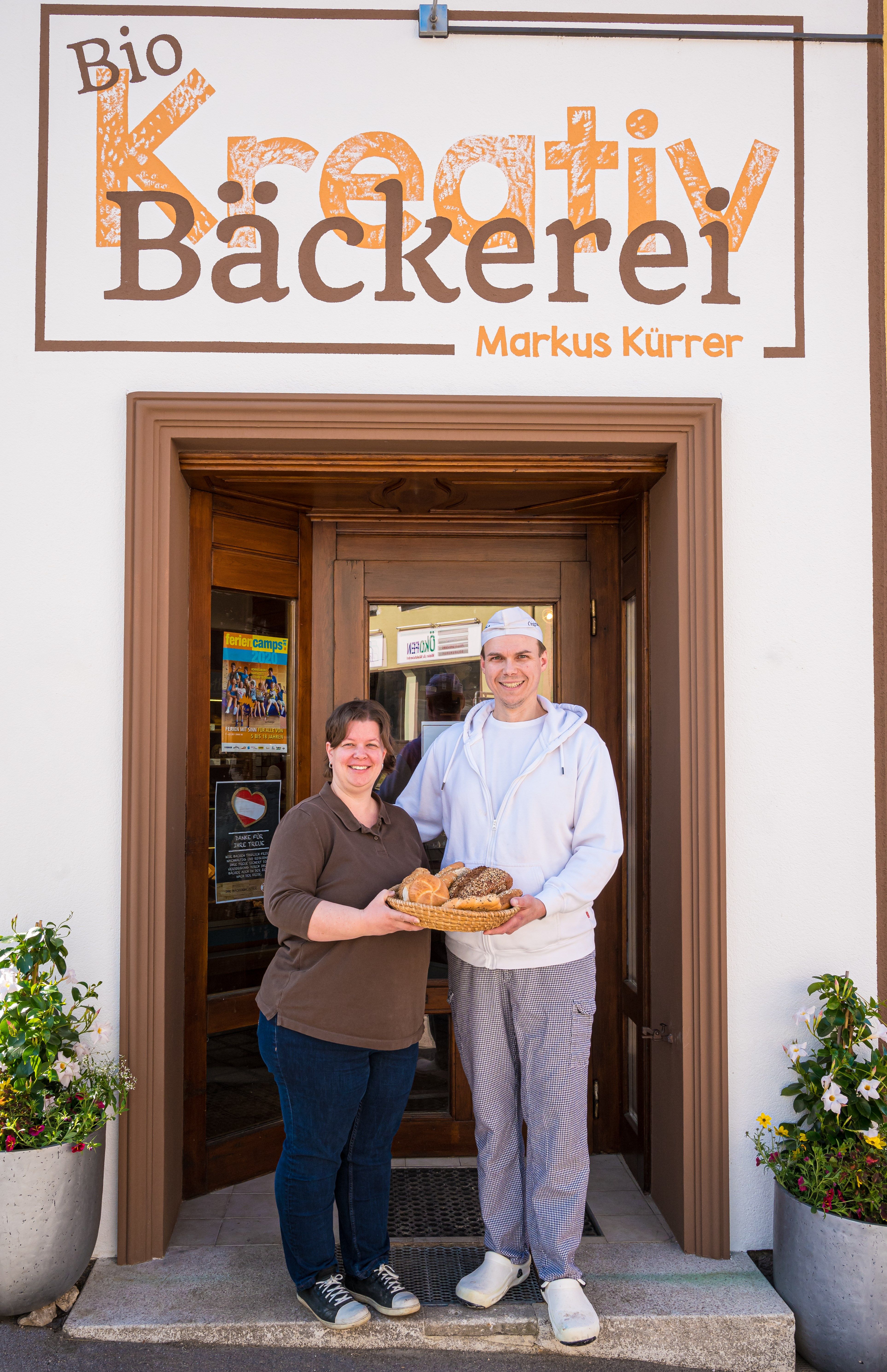 Zwei Personen stehen vor der Bio Kreativ Bäckerei Markus Kurrer und halten einen Korb mit Backwaren.