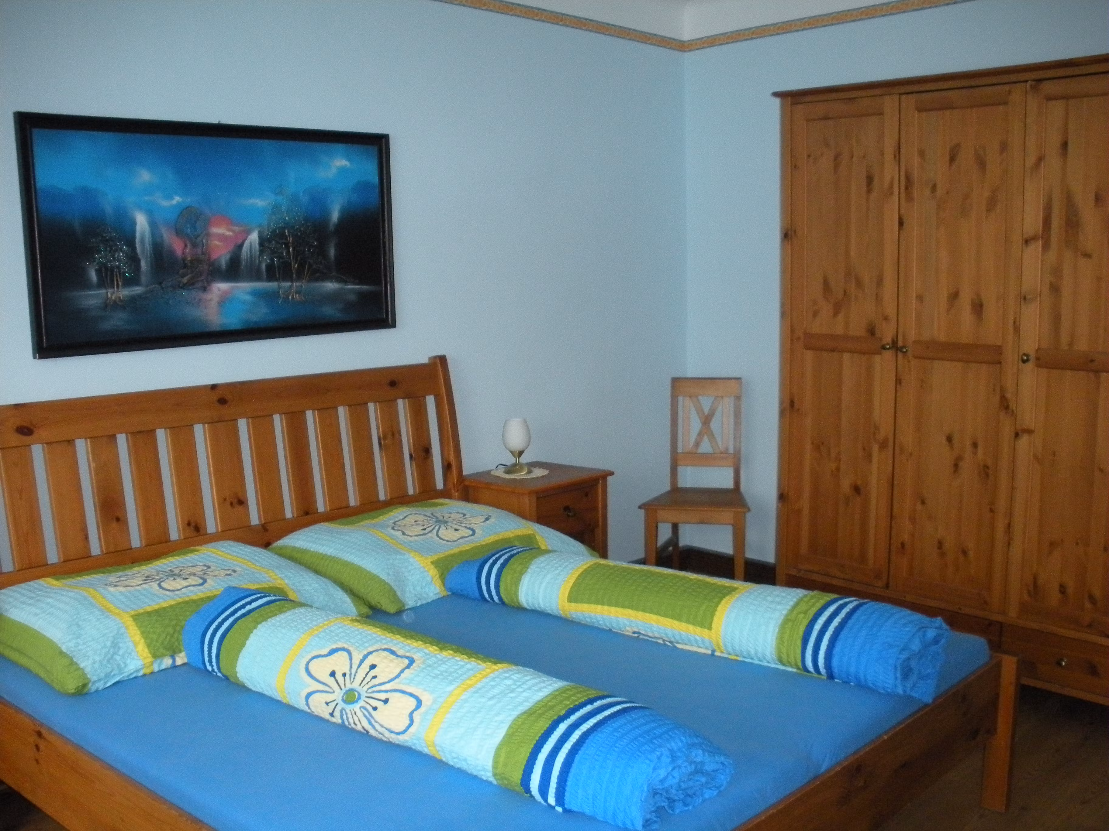 Ein Gästezimmer mit einem Holzbett, bunten Bettdecken, einem Schrank und einem Bild an der Wand.