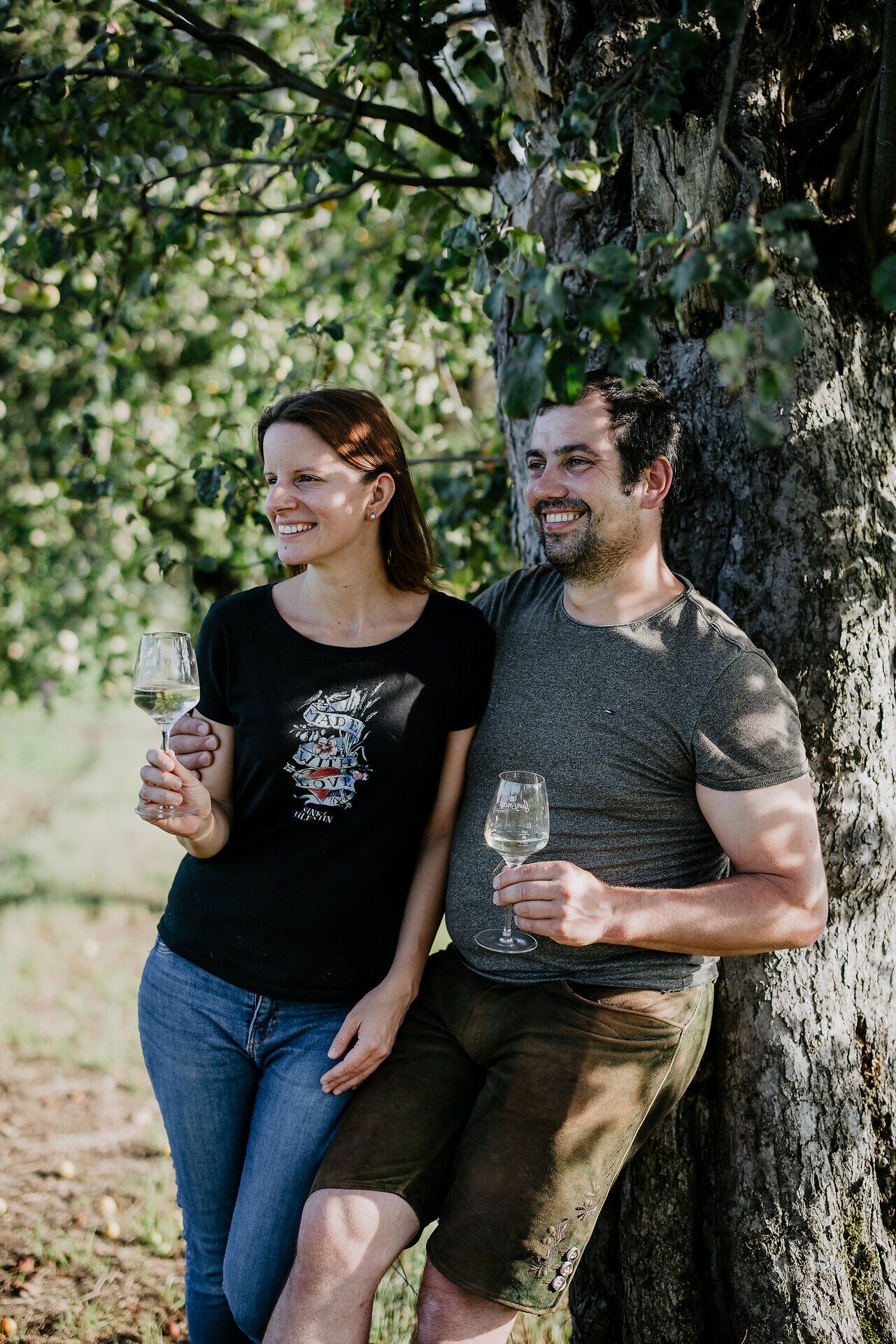 Inmitten der malerischen Landschaft des Mostviertels genießen Bernadette und Peter Haselberger einen entspannten Moment unter einem alten Baum. Mit Gläsern gefüllt mit köstlichem Birnenmost strahlen sie Freude und Gastfreundschaft aus, während die Sonne sanft durch das Blätterdach scheint.