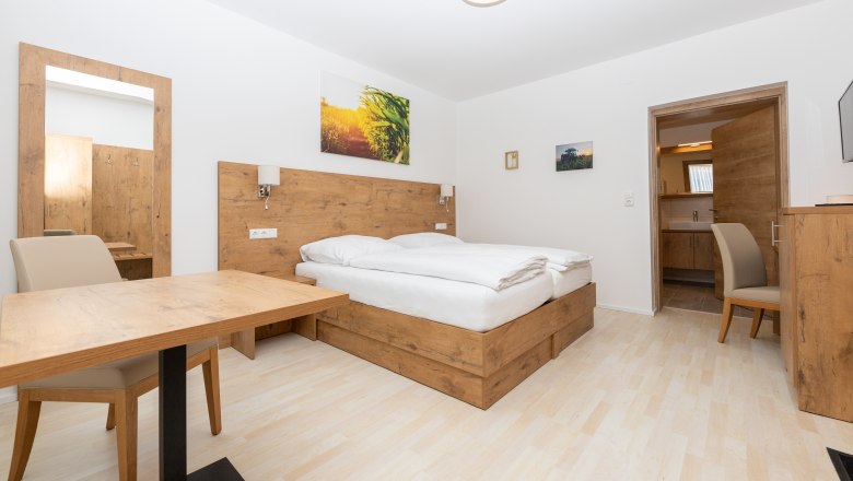 Schlafzimmer, © Schönfelderhof Modernes Schlafzimmer mit Holzmöbeln, Doppelbett, Tisch und Stuhl. Ein Spiegel und Bilder an der Wand, Tür führt zu einem Badezimmer.